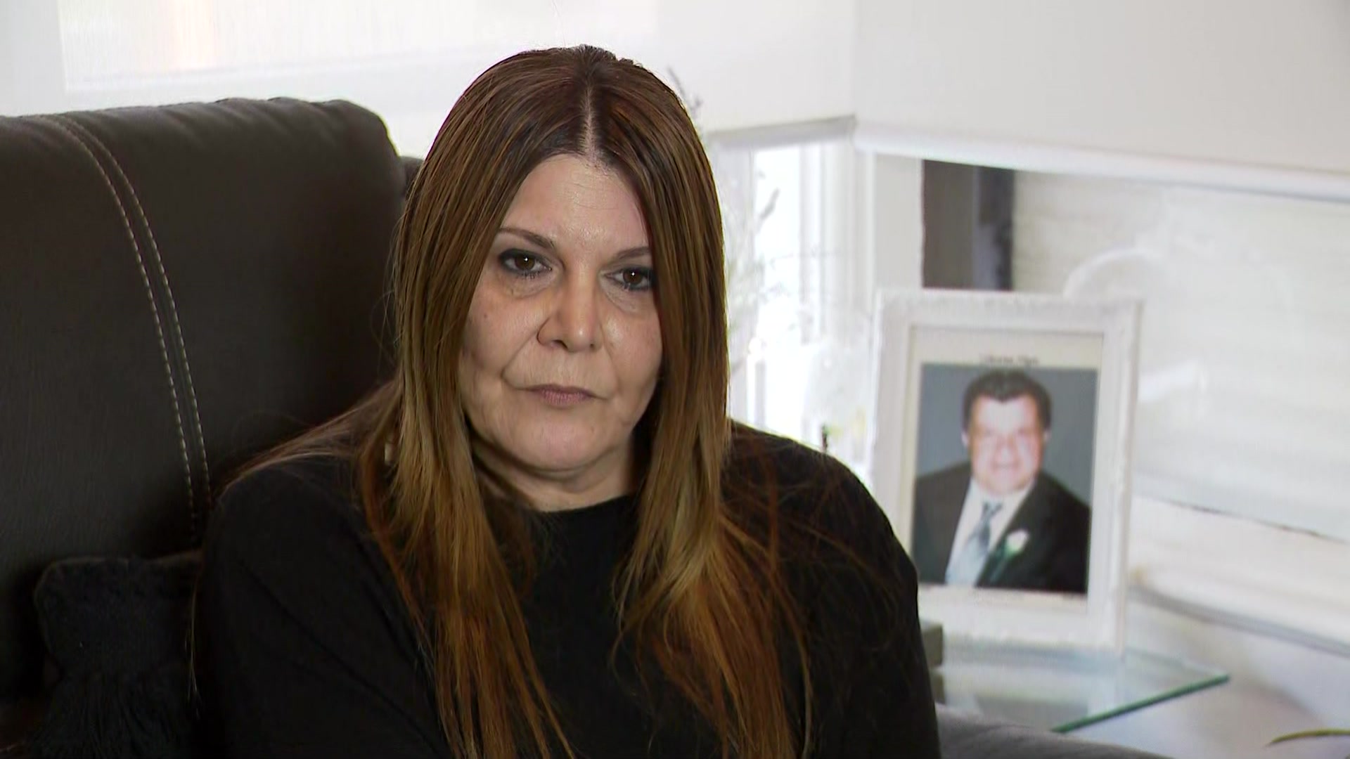 A woman sits on a couch, with a framed photo of a man behind her.