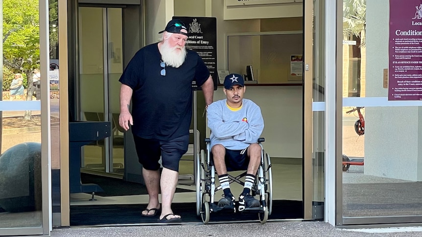 A young Indigenous man in a wheelchair, being wheeled out by an older man through the front of a court building's front door