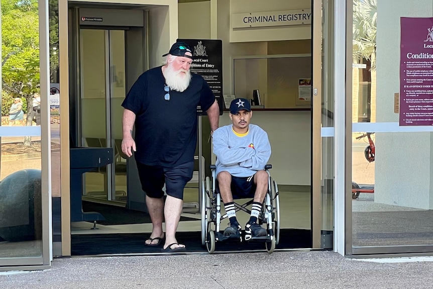 A young Indigenous man in a wheelchair, being wheeled out by an older man through the front of a court building's front door