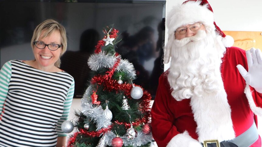 a lady in a white and blue striped top smiles next to a green Christmas tree and Santa Claus