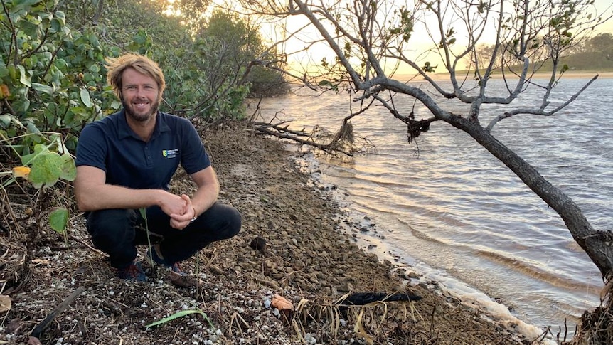 A man in jeans and a polo shirt smiles as he kneels beside a creek.