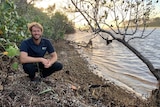 A man in jeans and a polo shirt smiles as he kneels beside a creek.