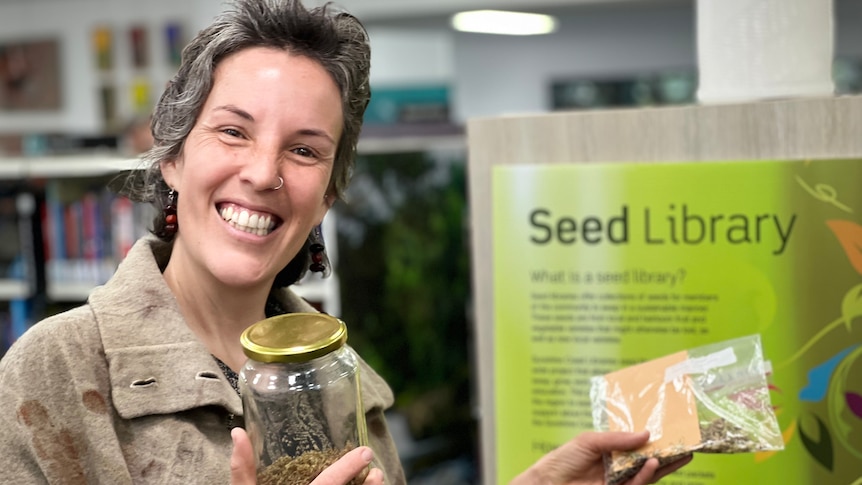 A smiling woman with short brown hair and a nose ring holds a jar with seeds isnide inside it.