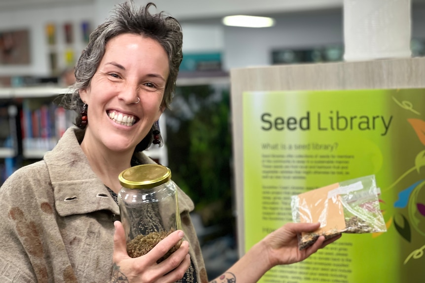 A smiling woman with short brown hair and a nose ring holds a jar with seeds isnide inside it.