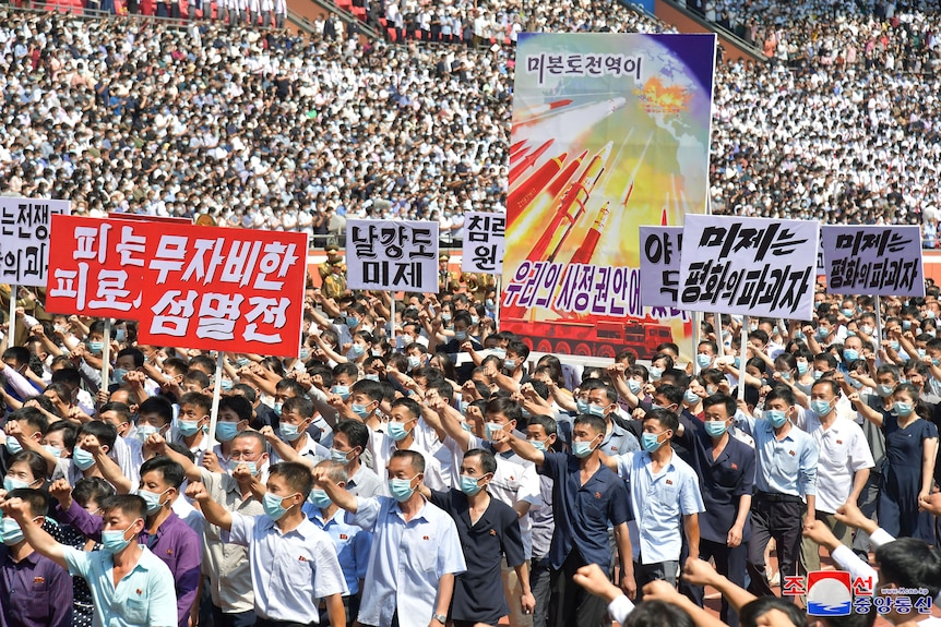 A crowd, mostly wearing face masks and raising one hand in the air, walk as part of a rally inside a stadium. Crowds in stand