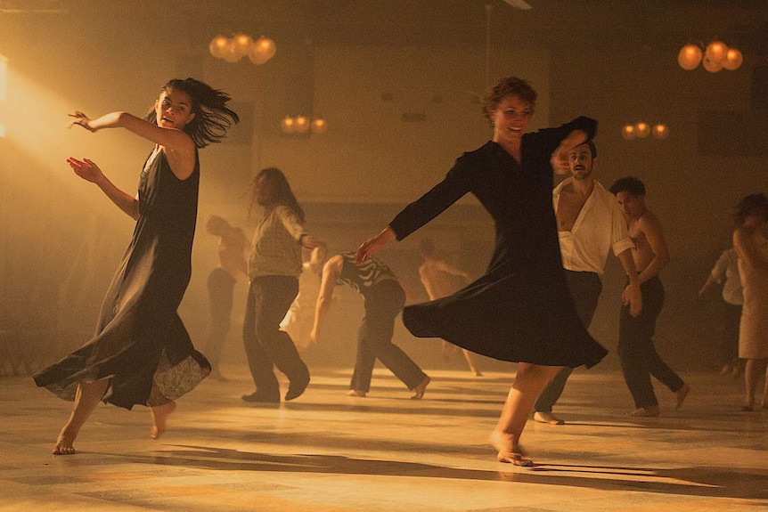 Many men and women dancing in a room lit by afternoon sunlight and dim overhead lamps.