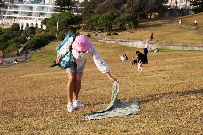 A woman wearing a pink hat inspects an abandoned towel on a grassy hill