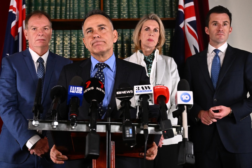 Victorian Opposition Leader John Pesutto stands at a podium with media microphones, with three colleagues standing behind him.