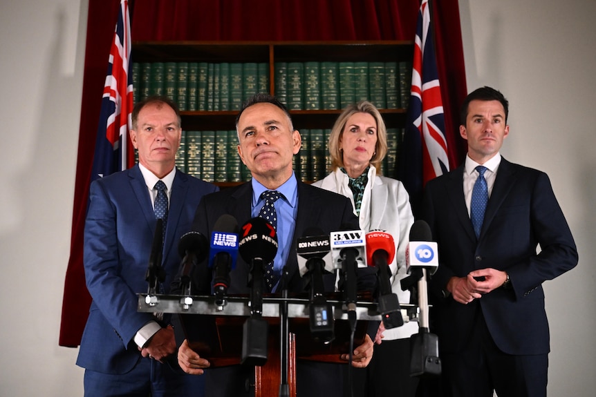 Victorian Opposition Leader John Pesutto stands at a podium with media microphones, with three colleagues standing behind him.