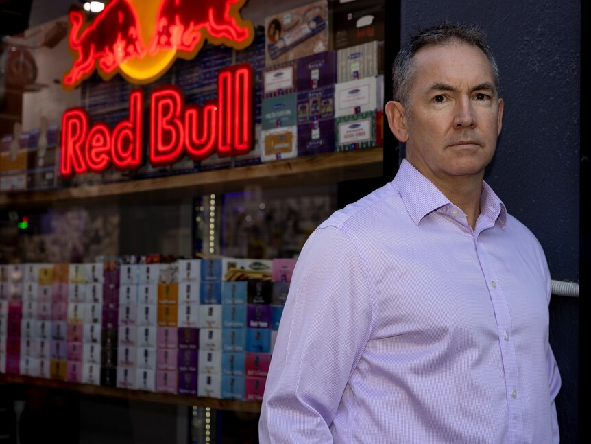 Man wearing blue shirt standing in front of a convenience store with a Red Bull logo behind him.