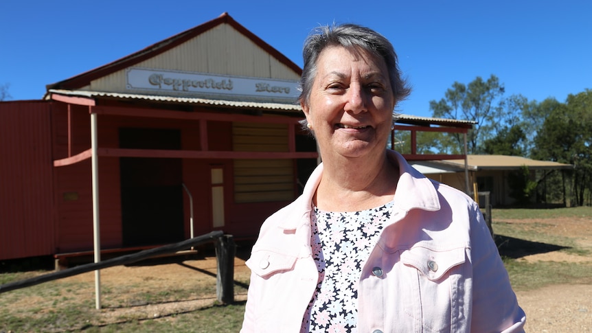 A woman in a white denim jacket stands smiling in the afternoon sun in front of a weatherboard and tin building from the 1800s.