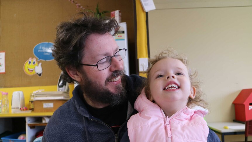 Bearded man with glasses smiling with his young daughter on his lap.