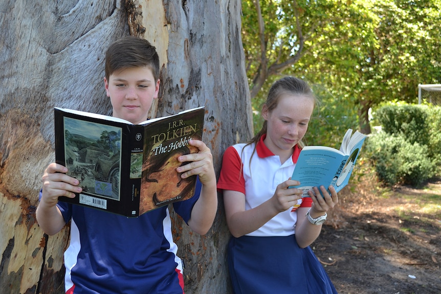 Colour photo of primary school students Harry and Annabelle standing and reading outside in front of tree.