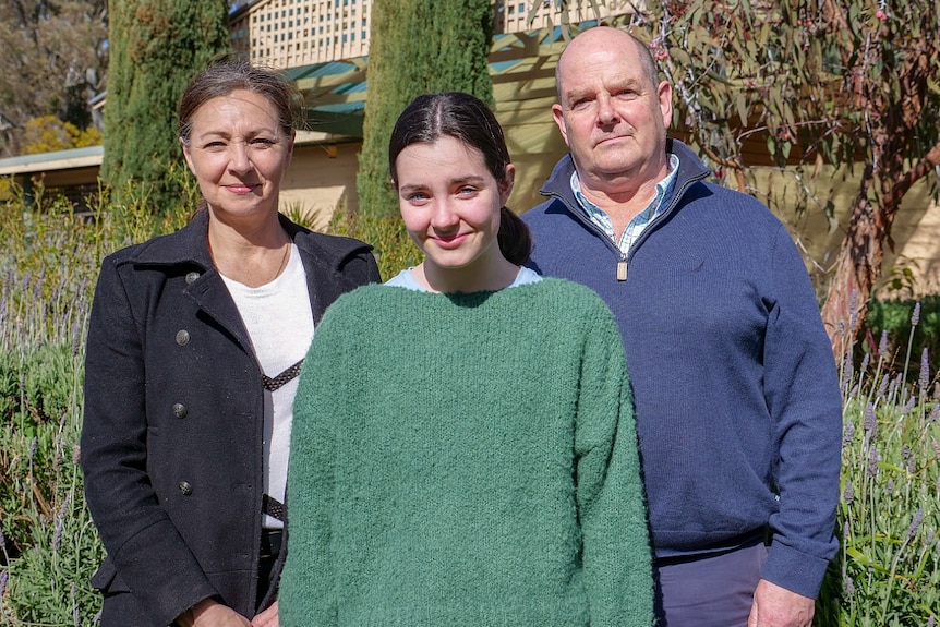 A man in a blue jumper and a woman wearing a dark jacket stand either side of a young woman in a green top.