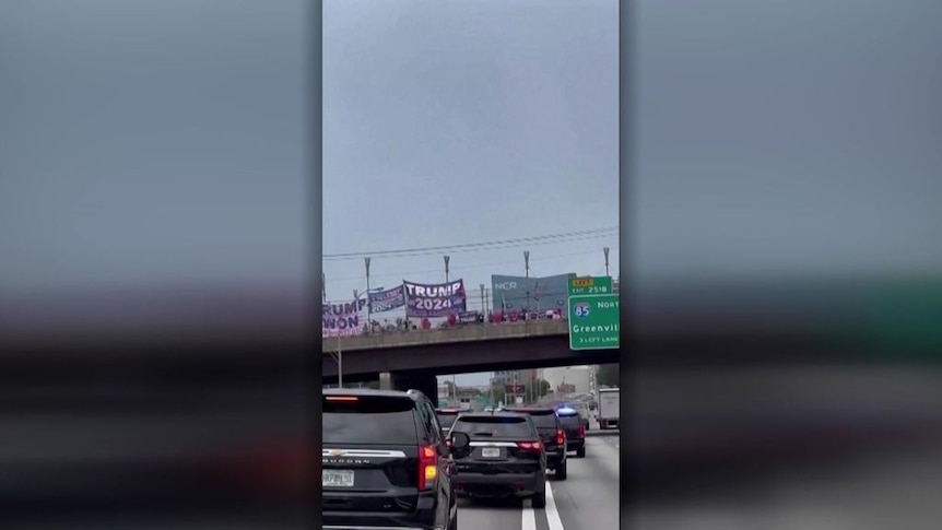 Screengrab of mobile vision showing banners and people on an overpass and a motorcade of black cars going underneath.