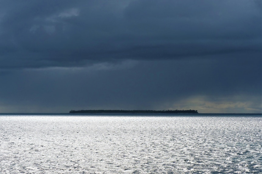 An island in the distance under a dark cloudy sky. The ocean in front is bright with the sun's reflection.
