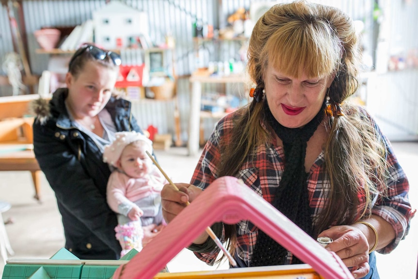 A woman paints doll house furniture as a younger woman with a baby watches on