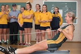a woman in rowing uniform goes through her paces on a rowing machine while students watch on
