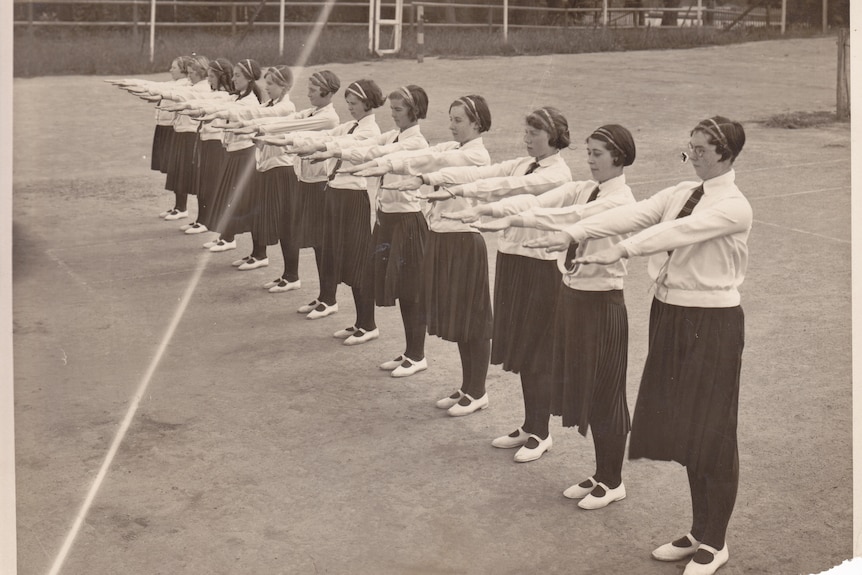 A black and white image of a line of women wearing skirts and long sleeve tops holding their hands out.