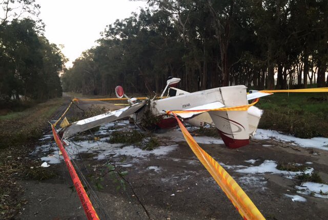 The wreckage of a white light plane sits upside down on a dirt road with trees lined up on either side.