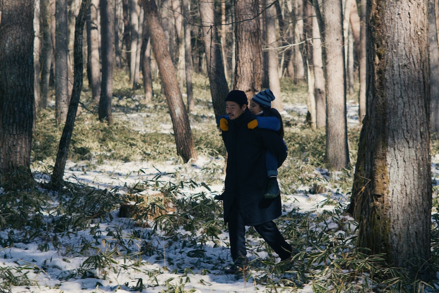 A girl piggybacks on a man's shoutlders as they walk through a winter forest, small amounts of snow in pockets between green.