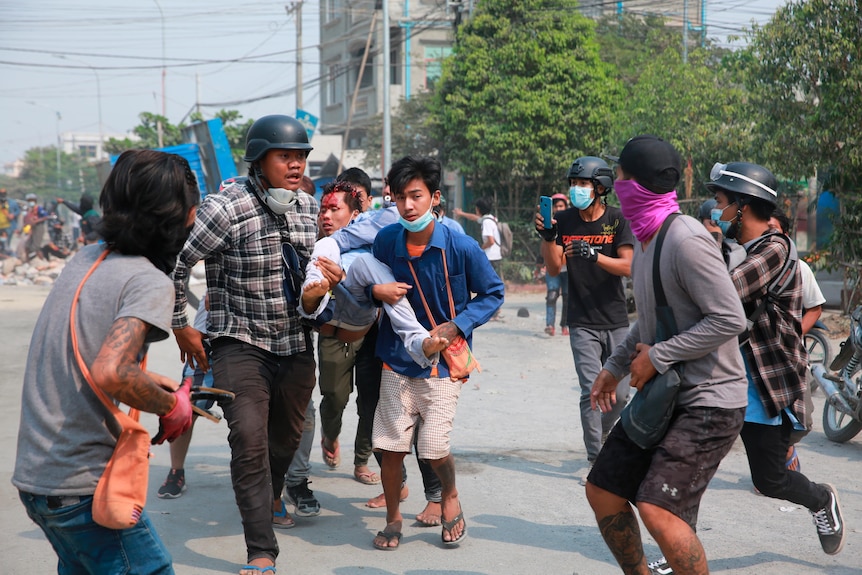 A man with a head injury is carried by other men running through the street.