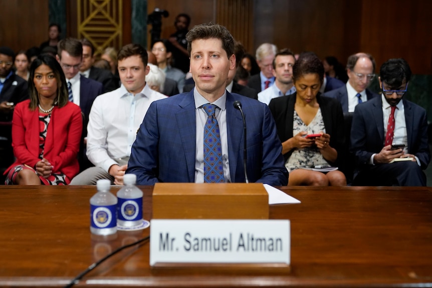 A man in blue suit sitting at desk with people sitting behind him. He has a straight face.