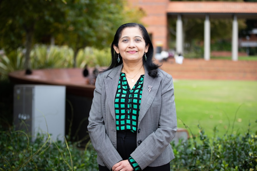 A woman in a gray blazer stands in front of a manicured lawn and manicured bushes.