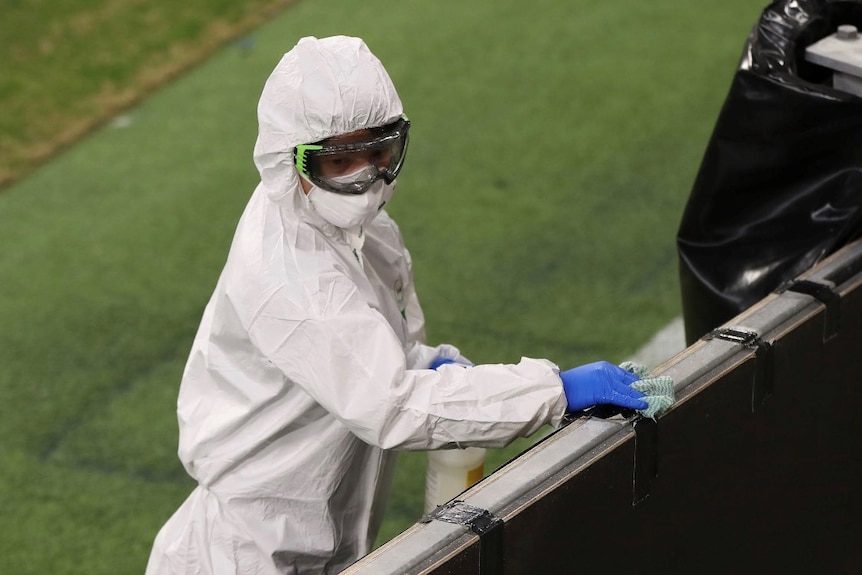 A cleaner wearing a white jumpsuits, face mask and goggles cleans the fence at Perth Stadium with a cloth.