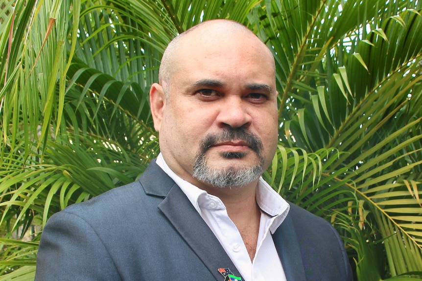 a bald man with facial hair wearing a suit in front of palm fronds