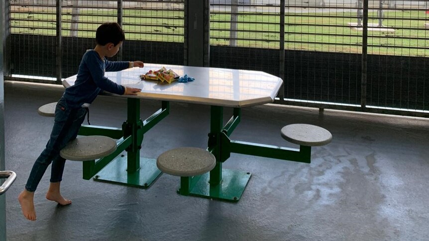 A boy in pyjamas leans over a table to select a treat with large fencing looming around him.