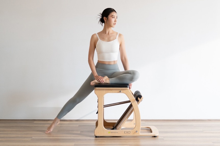 A young Asian woman sitting on a chair in a Pilates stretch.