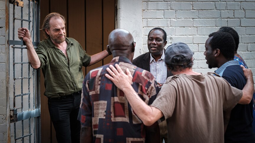 Colour still still of Hugo Weaving standing in doorway next to a small circle of men singing in 2019 film Hearts and Bones.