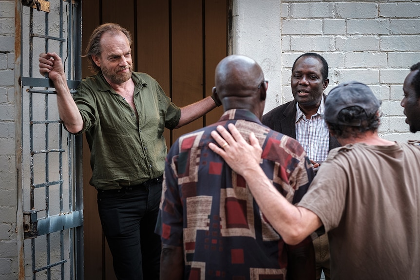 Colour still still of Hugo Weaving standing in doorway next to a small circle of men singing in 2019 film Hearts and Bones.