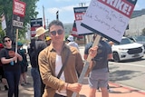David Koutsouridis with a sign, wearing sunglasses, people holding signs behind him at the writers strike picket lines