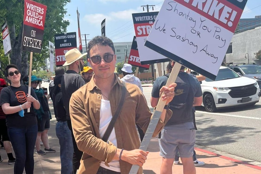 David Koutsouridis with a sign, wearing sunglasses, people holding signs behind him at the writers strike picket lines
