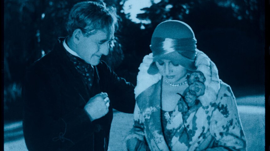 A blue-tinted image from a 1929 film showing an older man in a robe with his hand on a well-dressed woman's shoulder.
