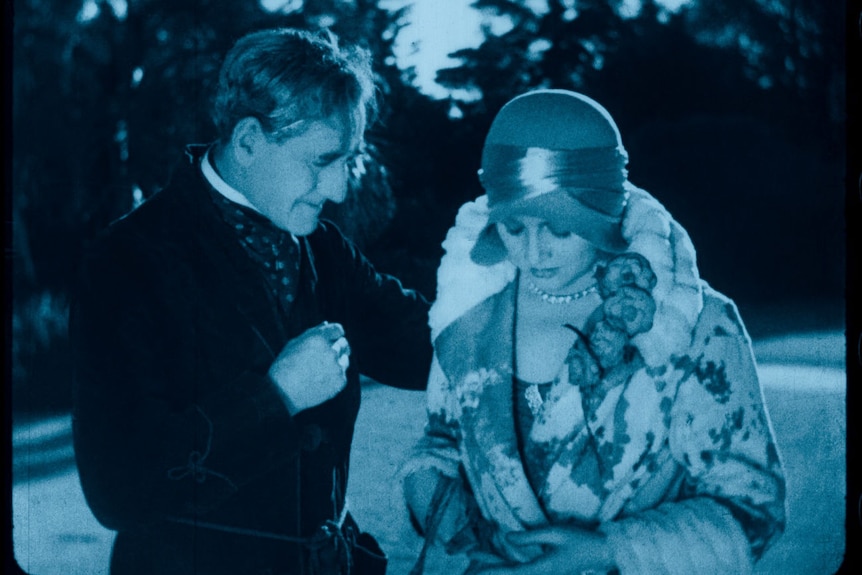 A blue-tinted image from a 1929 film showing an older man in a robe with his hand on a well-dressed woman's shoulder.