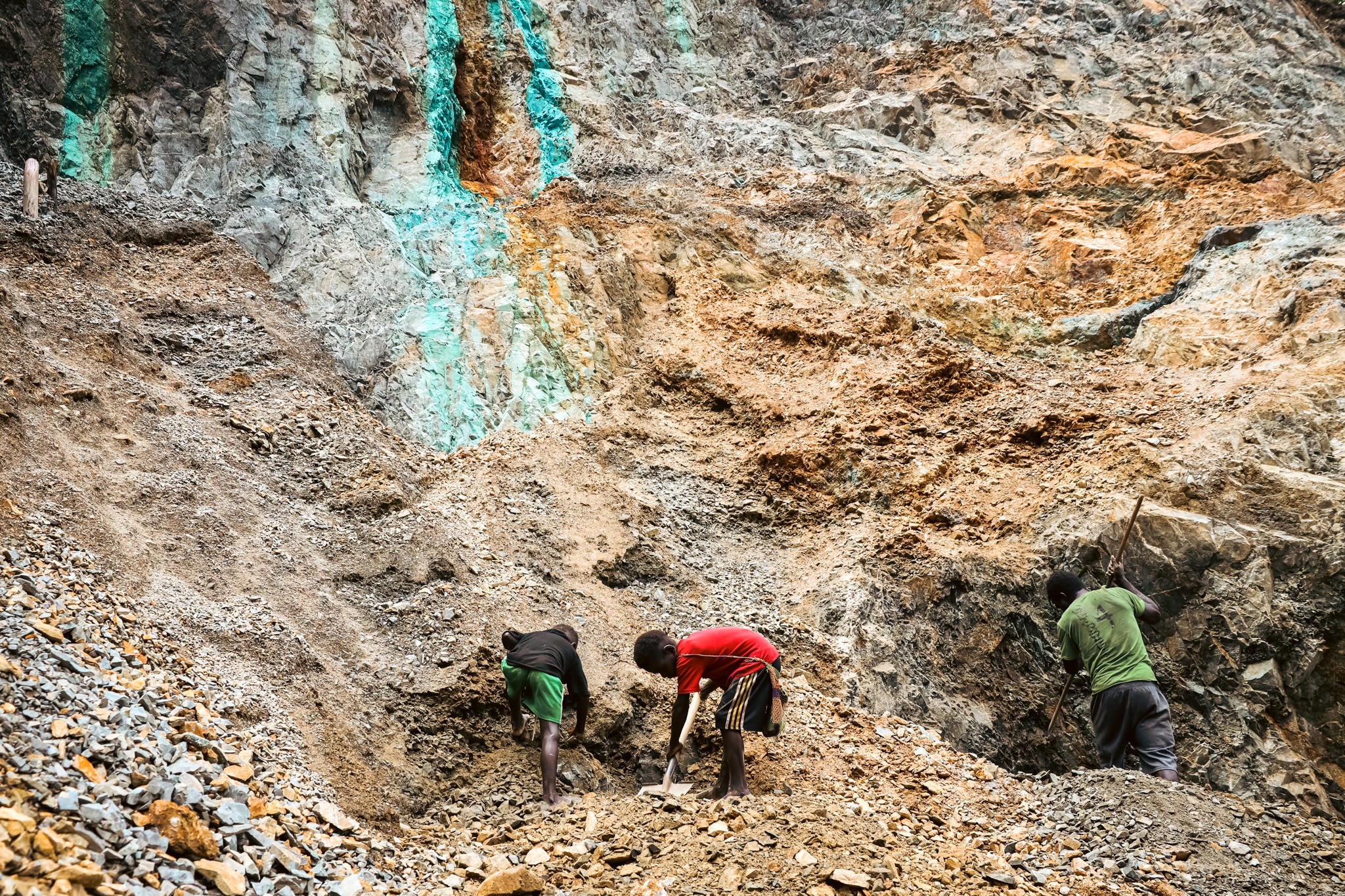 Three kids dig holes with shovels at the bottom of a mound of dirt.