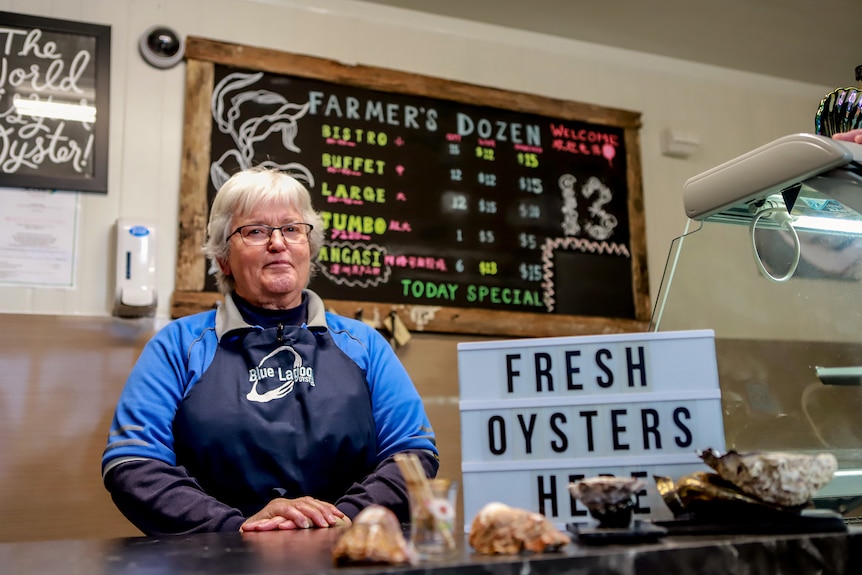 Woman with white hair wearing navy blue apron and blue shirt stands at shop counter 