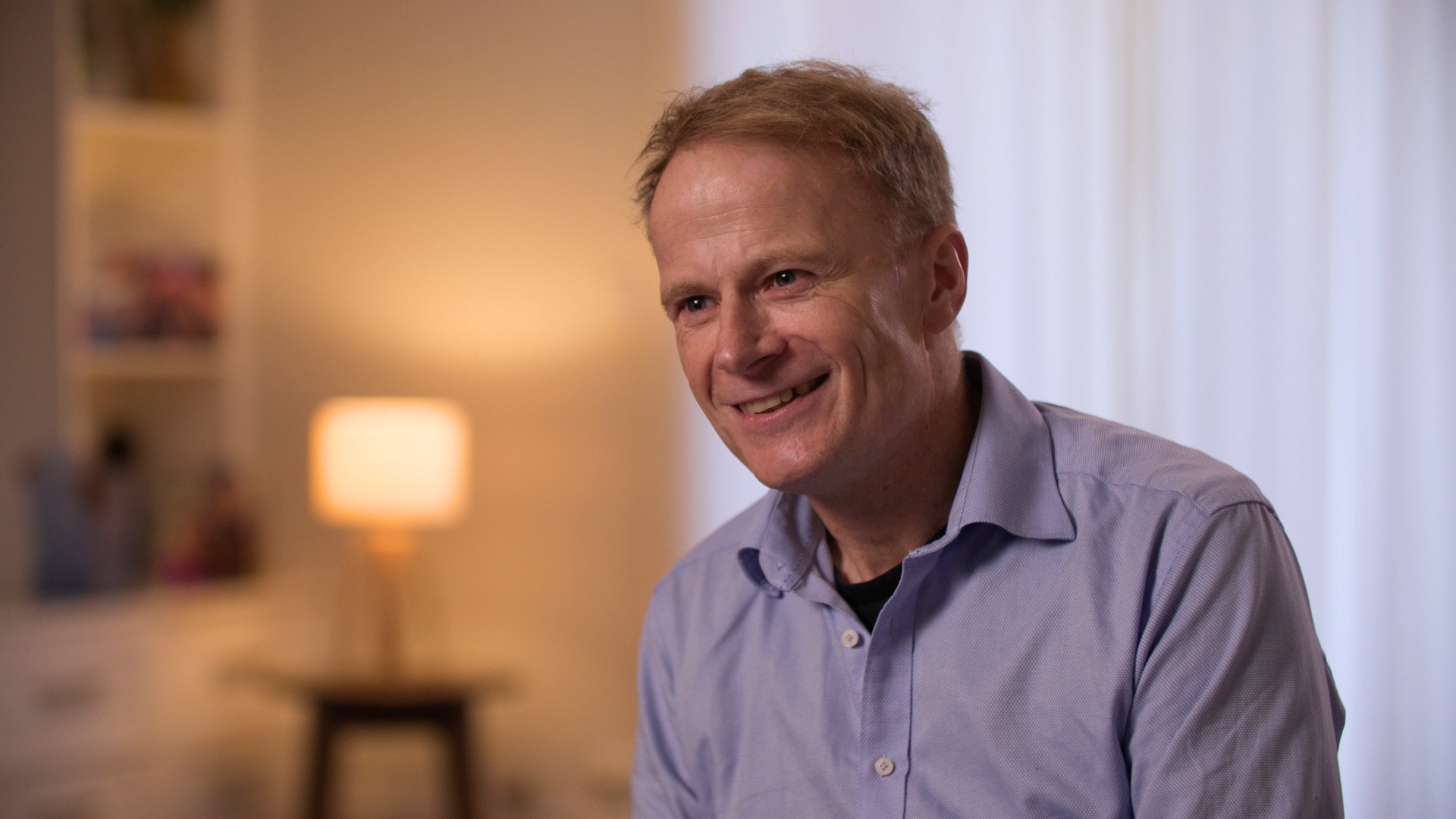A man with wispy short blonde hair wears a blue collared shirt, seated, he is smiling. A lamp on behind him
