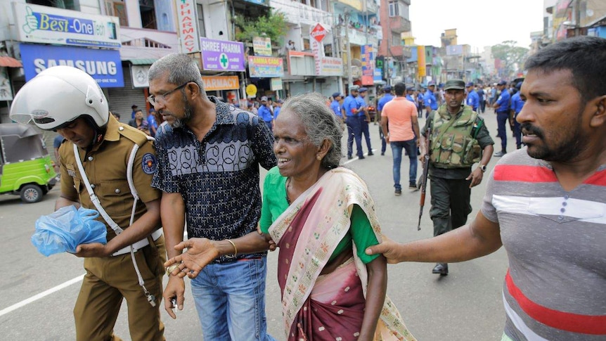 Three men walk with a distressed-looking elderly woman on a street with military personnel int he background.