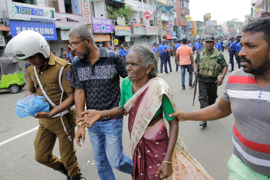 Three men walk with a distressed-looking elderly woman on a street with military personnel int he background.