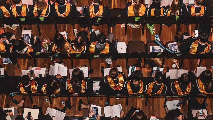 An aerial view of rows of seated graduation students with colourful sashes over their black academic robes.
