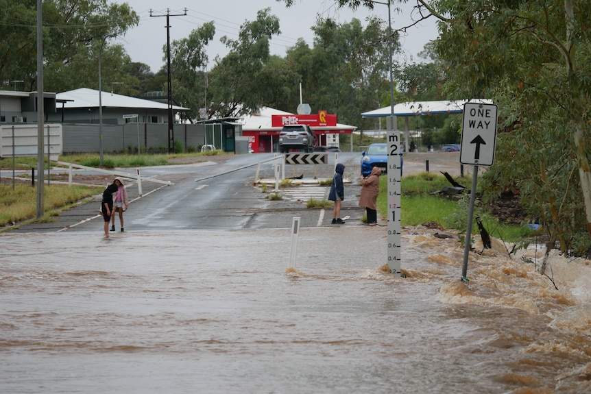 People stand on the far side of a river bank, looking cold on a grey day as the brown water cuts off a road.