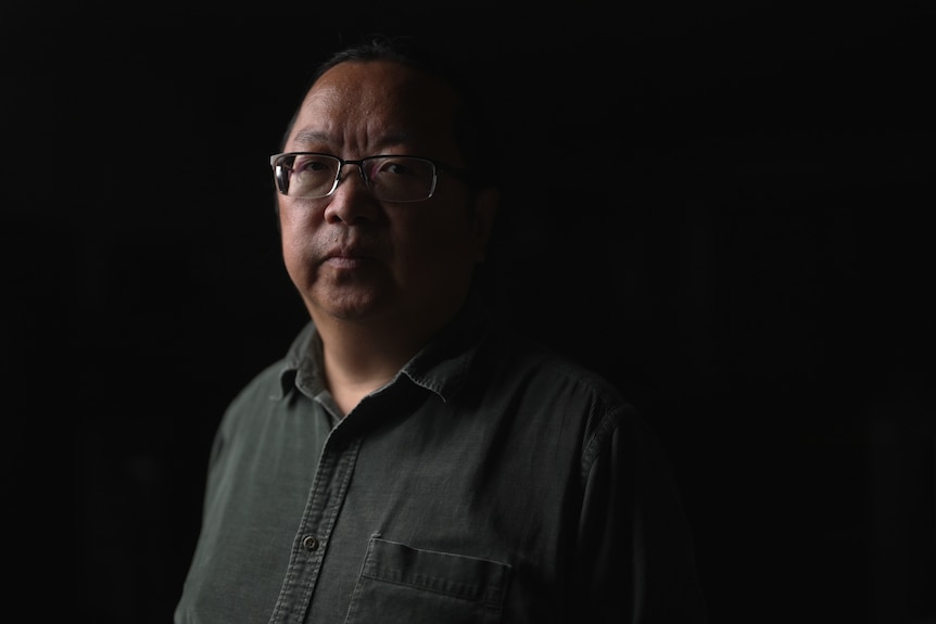 A man in a dark shirt and glasses stands in a darkened room.