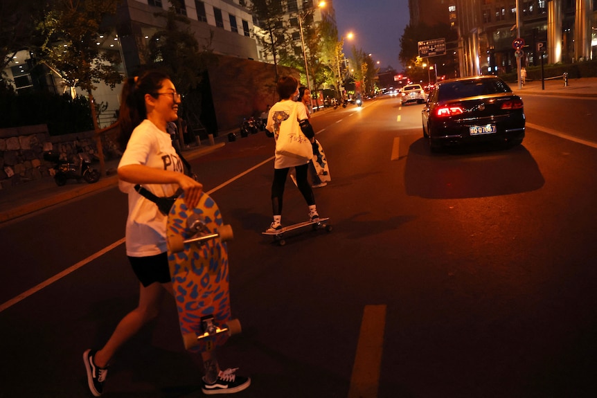 One woman carries her skateboard and another is on a skateboard on a busy street at night. 