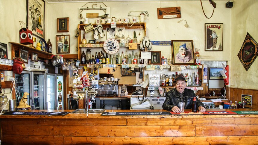 A man in a black leather jacket behind the bar, an empty beer glass and beer bottle on the bar beside him.