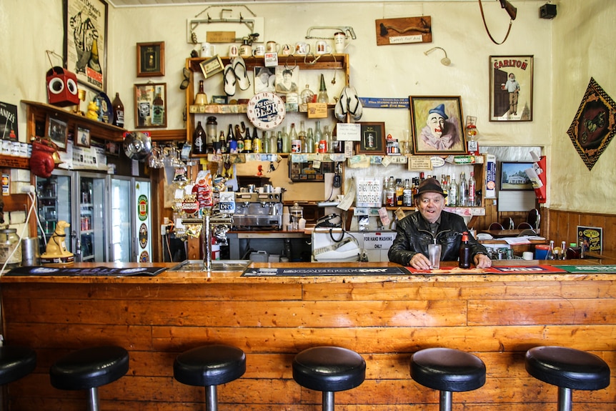 A man in a black leather jacket behind the bar, an empty beer glass and beer bottle on the bar beside him.