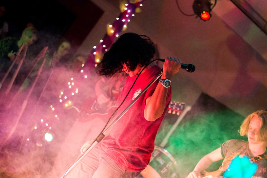 A dark haired boy holds a microphone on a stage covered with smoke. There are pink and green lights.